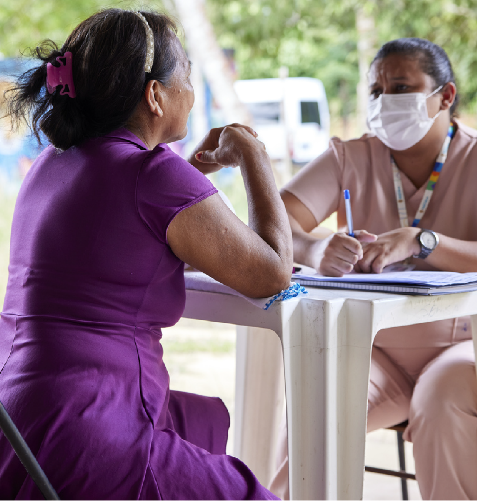 Woman in purple dress sitting across the table from a woman in pink scrubs and a mask.