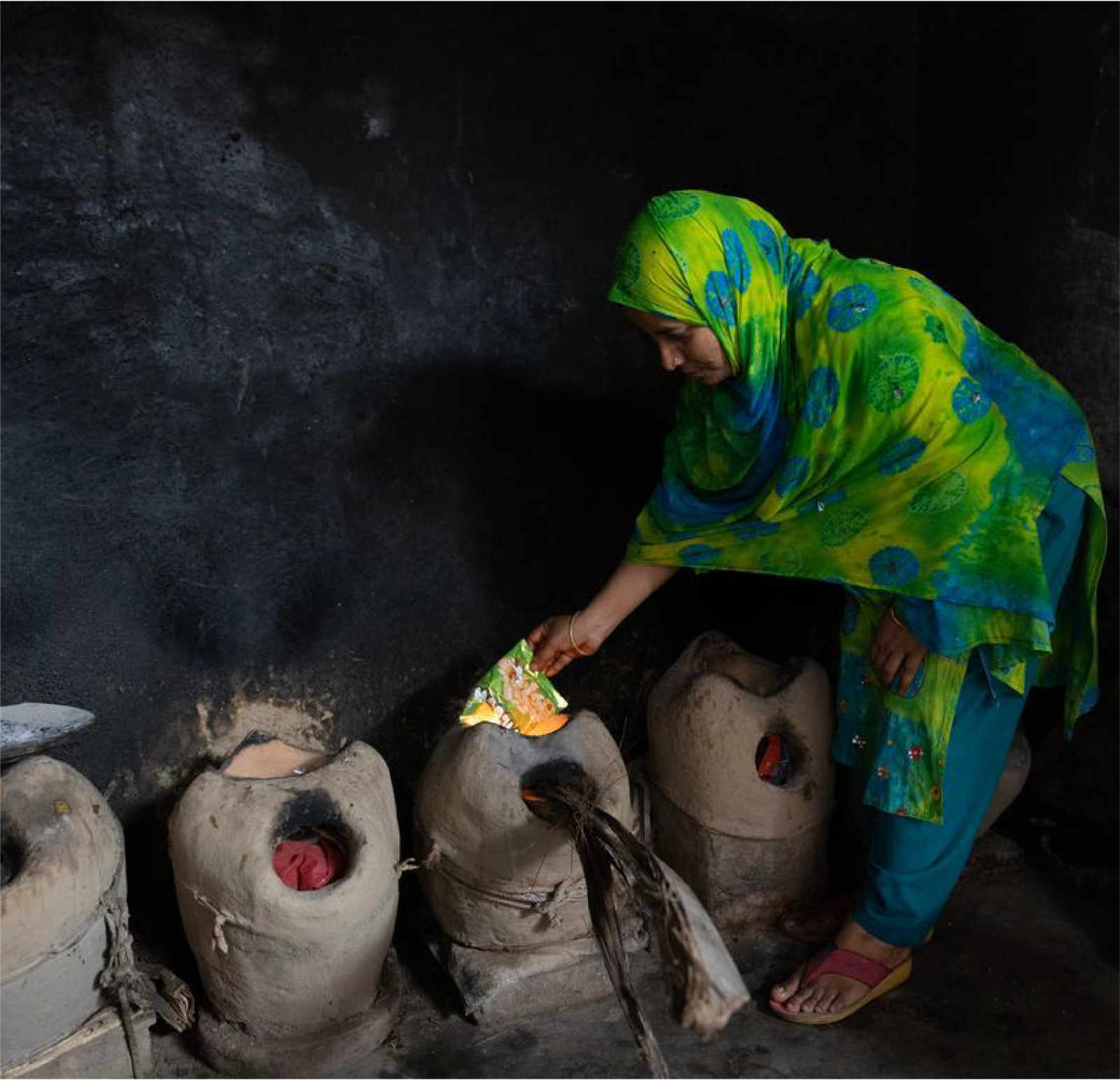 Woman in green and blue sari cooking inside over fire pits.