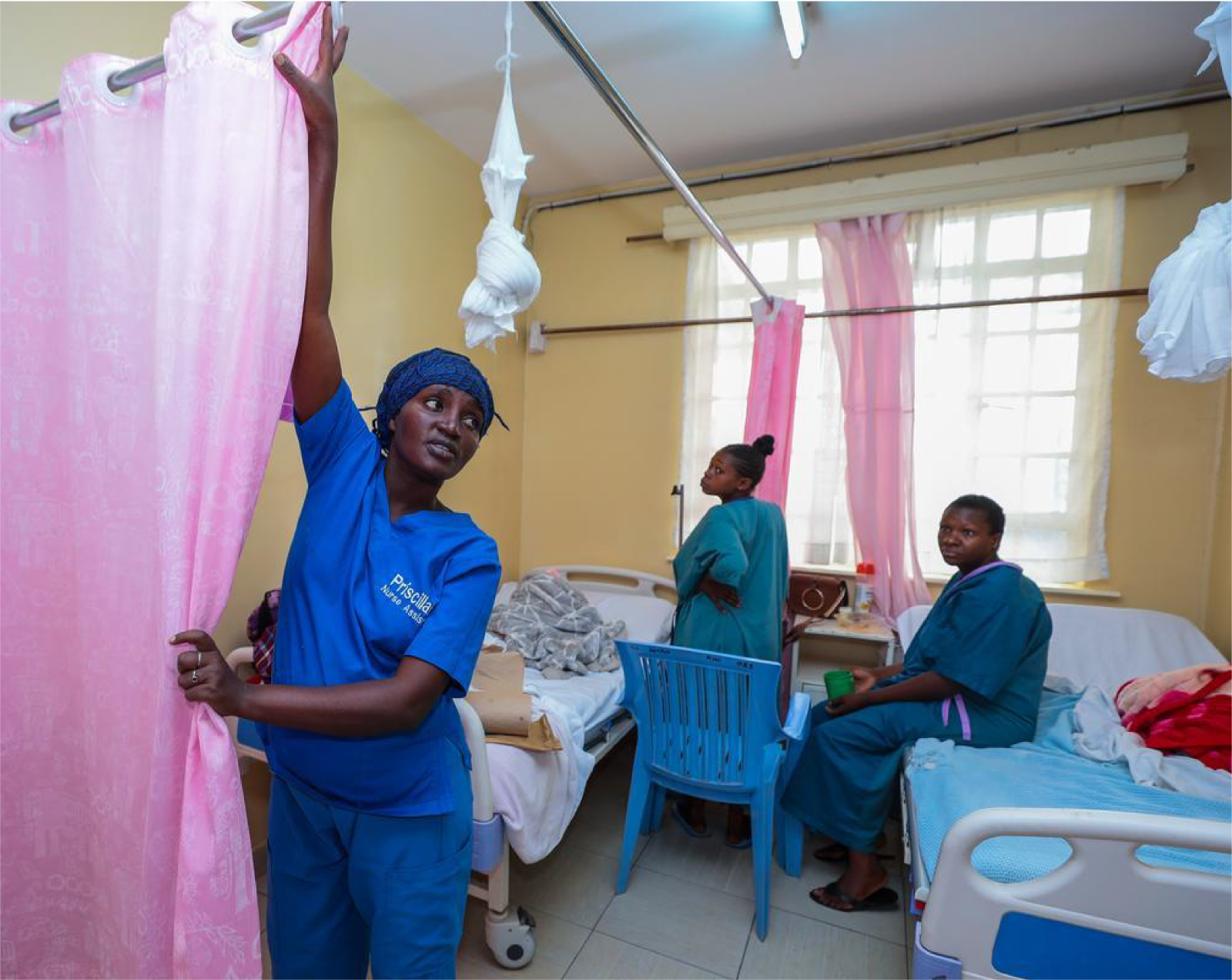 Kenyan nurse dressed in blue scrubs closing a pink curtain to maintain privacy for two other woman patients.