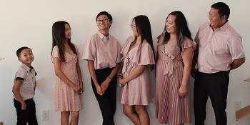 A family of six poses smiling together against a white wall, all dressed in matching pink outfits. Mother and father look at their elementary aged child and three adolescent children.