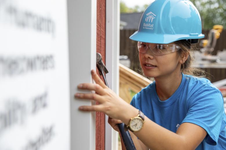 A young woman in a blue hard hat works on the siding of her future Habitat home.