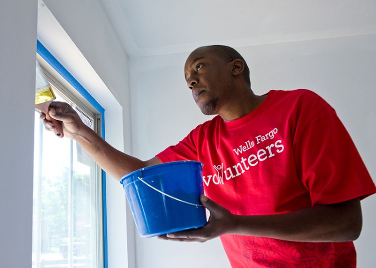 A man in a red t-shirt with "Wells Fargo Volunteers" on the front paints a window frame inside a Habitat house.