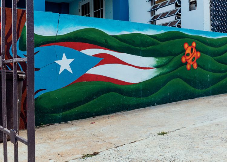 A mural on a concrete wall shows the Puerto Rico flag blending into a green mountains and blue sky.