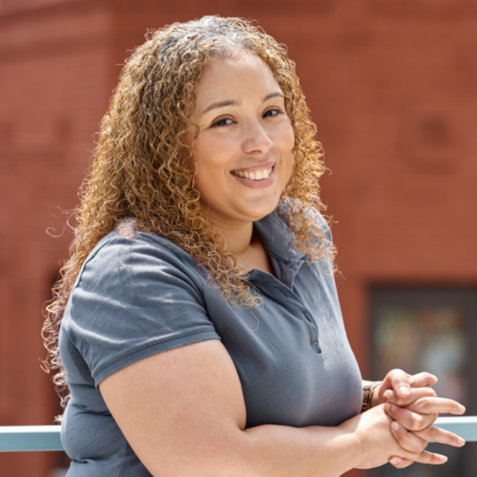 A headshot of Janet smiling in a grey shirt with a red-brown wall in the background.