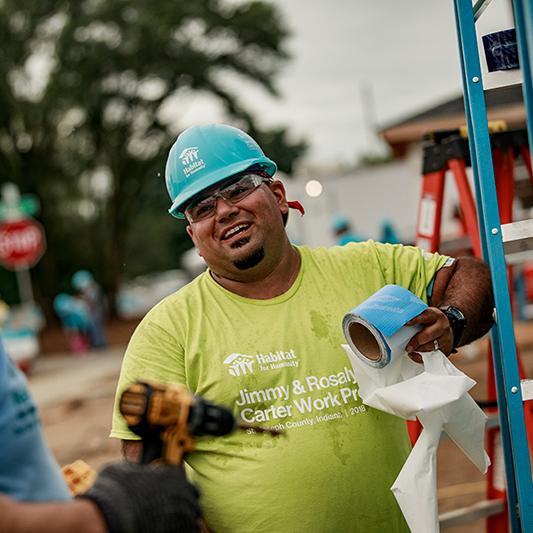 Benito in a green Habitat volunteer shirt and blue hardhat talking to another volunteer on a build site.