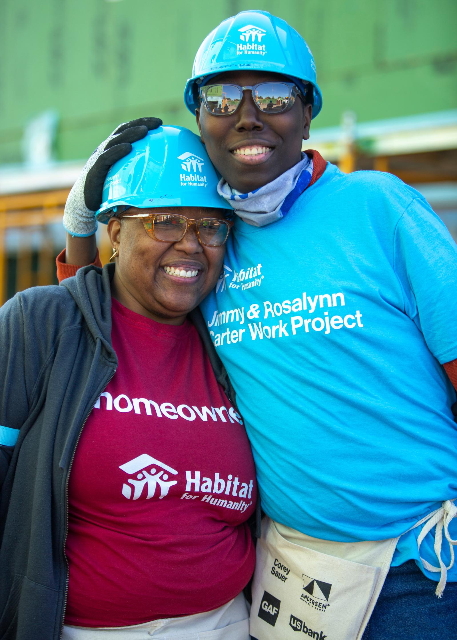 Buffy and her adult son in blue hardhats smiling at the camera on the buildsite.