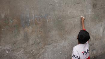 A child writes "Home" with chalk on a gray concrete wall.