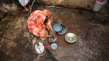 A woman in an orange dyed scarf sitting on the ground and cleaning plates and bowls.