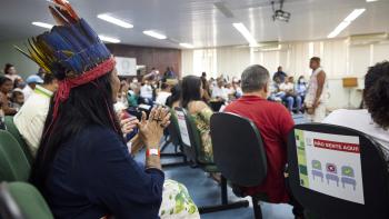 A woman in a traditional Brazilian headdress clapping her hands together at a community meeting. 