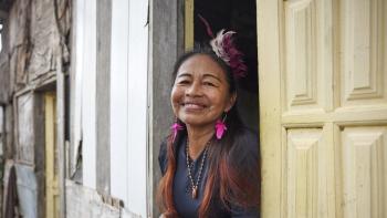 An older woman peers out from a window with pale yellow shutters, smiling.An older woman peers out from a window with pale yellow shutters, smiling.