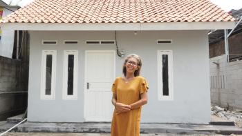 A woman in an orange shirt smiles in front of her home.