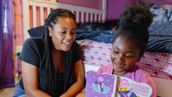 Young girl smiling while reading a picture book with her mother in her pink and purple bedroom.