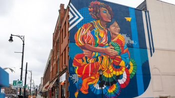 Mural entitled "Sisters" is painted on the side of a brick building and depicts a Black woman and a Latina woman embracing, painted in bright rainbow colors.