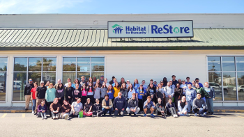 Large group of Collegiate Challenge student volunteers pose under Habitat ReStore sign