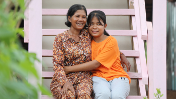 A Cambodian woman and her adolescent daughter hug and smile together while sitting on the pink wooden stairs of their Habitat home.