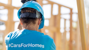 Habitat volunteer in blue hardhat and shirt that reads "#CarsForHomes" helps raise a wall on a build site.