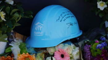 Laying on a bed of flowers is a light blue hardhat with the Habitat logo and a message written on the side that reads "Thank you for a lifetime of service and inspiration. We are forever grateful" signed by Habitat CEO Jonathan Reckford.