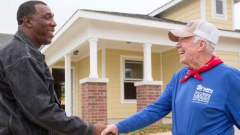 President Carter in red bandana and blue Habitat shirt shaking hands with man in front of Habitat house