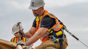 Man in construction gear and hard hat hammers nails on top of a roof