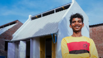 Man in a yellow shirt smiling and standing outside a white house.