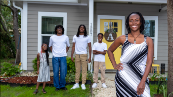 In front of her grey Habitat house with a decorated yellow door, a Black woman smiles with her three sons and young daughter behind her.