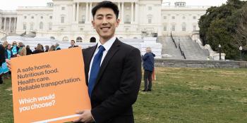 Man in suit standing in front of U.S. Capitol building holding s-ign that says "A safe home, nutritious food, healthcare, reliable transportation: which would you choose?"