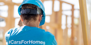 Habitat volunteer wearing blue hardhat and shirt that reads "#CarsForHomes" helps raise a wall on a build site.