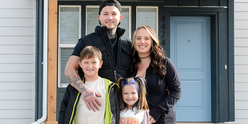 Young couple with two young children embrace and smile together in front of their Habitat home.