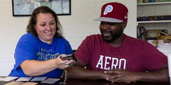 A woman and a man sit at a table with paperwork on it. The woman is showing the man something on a phone and they are both looking at it and smiling.