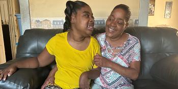 A mother and her young daughter, who has a disability that visibly affects her arms, are looking and smiling at each other while sitting on a couch together.