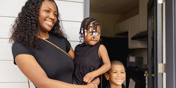 Mother smiles while holding her toddler-aged daughter and standing with her young son in the doorway of their Habitat home. 