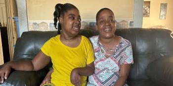 A mother sits on a couch smiling next to her young daughter who has a disability that visibly affects her eyes and arms.