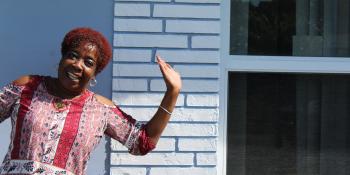 A woman posing joyfully with her hand in the air in front of a wall of white bricks.