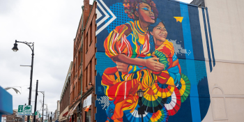 "Sisterhood" mural by Sylvia Lopez Chavez on a building in downtown Pittsfield depicts a Black woman and a Latina woman embracing, painted in vibrant rainbow colors.