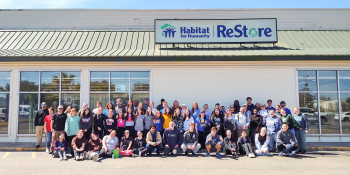 Large group of Collegiate Challenge volunteers pose together in front of Habitat ReStore sign.