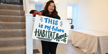 Woman inside a an empty room in a house holding a sign which reads "This is my future Habitat home"