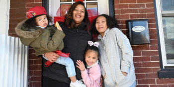 Mother smiling with three children by their home's front door with a big red bow on it.