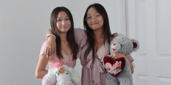 Two teenaged girls pose smiling together with stuffed animals in their home. 