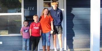 Mother and three children stand together on the front porch of their home.