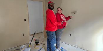 A couple wearing red sweatshirts smiles at the camera while painting the walls of their Habitat home.