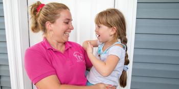 Mother in pink shirt holding young daughter in front of their home.