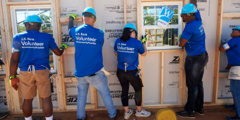 Five people wearing hard hats and blue "U.S. Bank volunteer" shirts work together to raise a wall on a Habitat build site