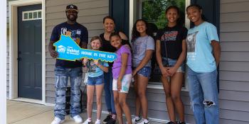 Family of seven smiling together on front porch, holding large key-shaped sign featuring Habitat logo and the phrase "Home is the Key" 