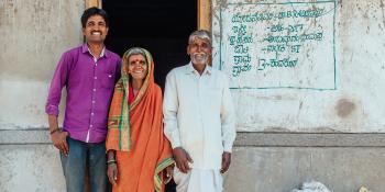Indian family of three in colorful clothing stand in front of their concrete home.