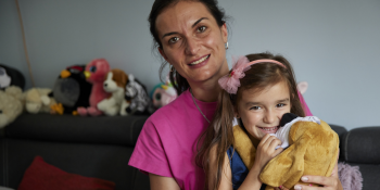 Young girl smiles as she hugs a stuffed puppy toy while sitting with her mother on their couch. 