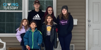 Mother with two teenagers and three young children pose together next to the front door of their Habitat home. Giving Tuesday logo is on the corner of the image.