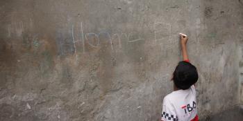 A young boy writes "home" in chalk on a cement wall.