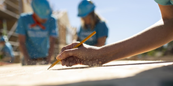 A close-up photo of a hand at a drawing board with two volunteers in blue shirts and hard hats blurred out in the background.