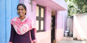 A woman stands outside a home painted pink, smiling.