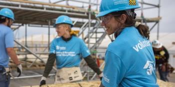 Two women in Habitat volunteer shirts carry a sheet of plywood on the 2024 Carter Work Project build site.